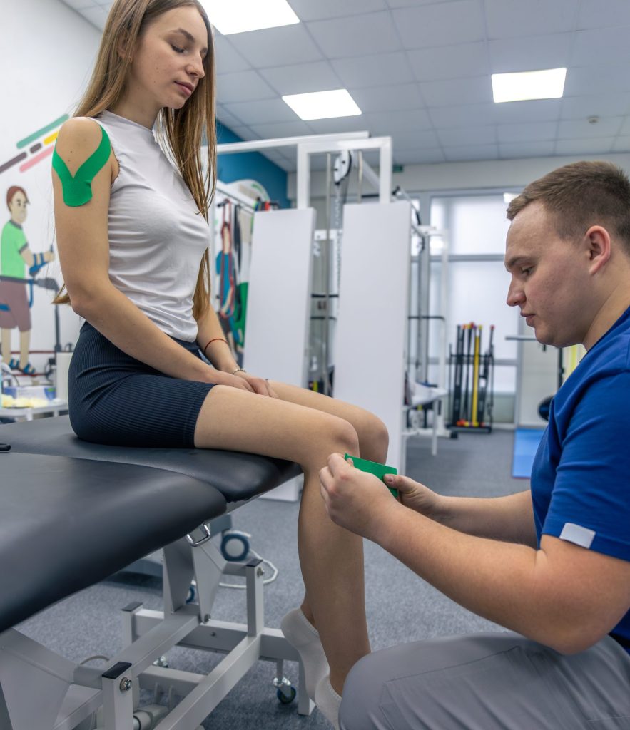 Physiotherapist applying kinesiology tape to a woman’s knee while she sits on a treatment table in a rehabilitation clinic.