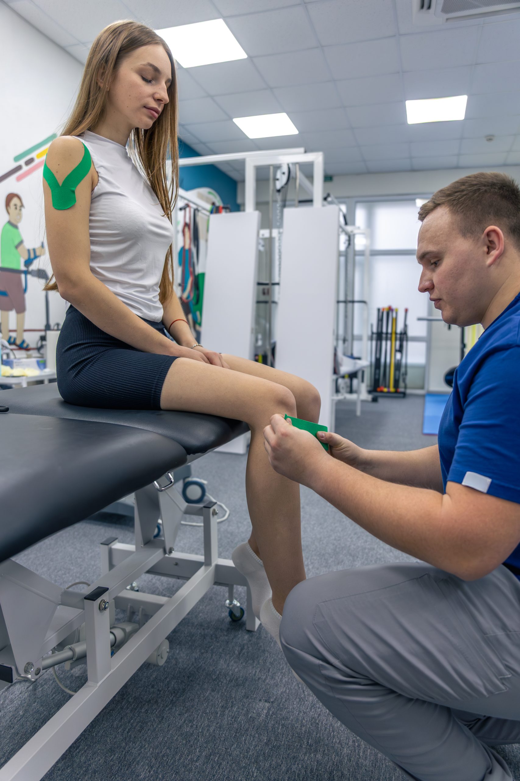 Physiotherapist applying kinesiology tape to a woman’s knee while she sits on a treatment table in a rehabilitation clinic.