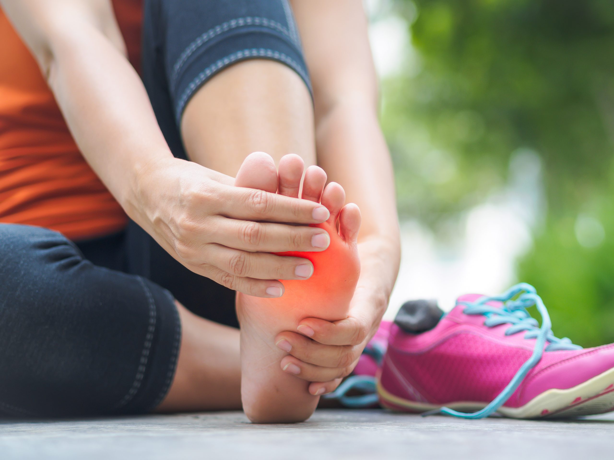 A person in workout clothes holds the ball of their bare foot, highlighted red to indicate pain, beside running shoes.